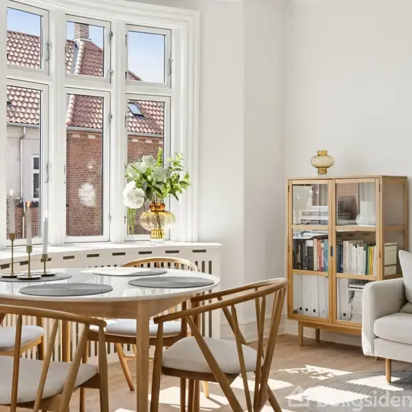 Rundt spisebord i lyst træ omringet af fire stole, står ved et stort, lysfyldt vindue. Ved siden af ses en sofa og en bogreol med glaslåger i et lyst rum.