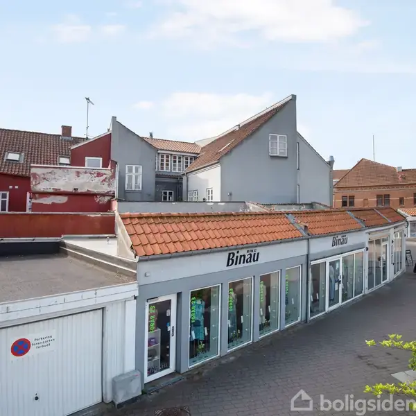 En butik med skiltet "Binäu" har store glasvinduer og ligger i en bygning med rødt tegltag. Omkringliggende er flere murstenshuse og en flisebelagt plads. En bænk står tæt ved.