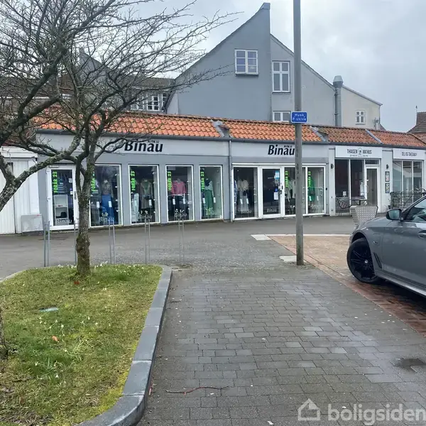 Butik med navnet "Binau" fremviser tøj i vinduerne. En grå bil holder parkeret i forgrunden. Træer og græsplæne omkranser fortovet i et bymiljø.