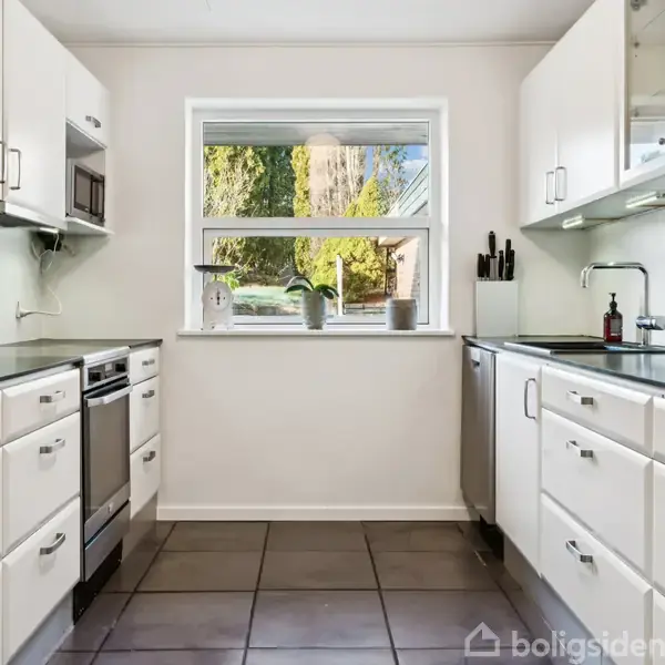 Køkken med hvide skabe på begge sider, stålkomfur og opvaskemaskine i moderne design. Et vindue i midten giver udsigt til en have, mens flisegulvet fuldender rummet.