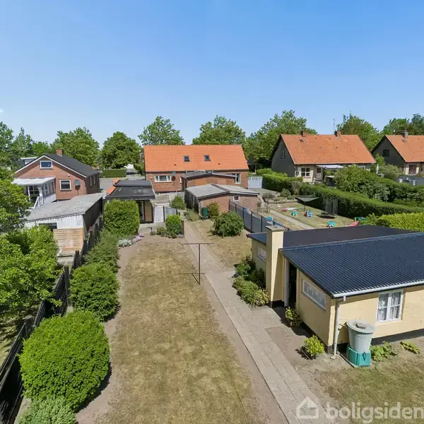 Hus med rødt tegltag står i en række af lignende huse. Foran huset en have med en flisebelagt sti og grønt område. Solrig dag med træer i baggrunden.