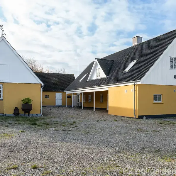 Gult landhus med sort tag står stille på en grusbelagt gårdsplads. Huset har gavltag, kviste og flere vinduer. En lille havekrukke står nær hoveddøren. Himmel med lette skyer.