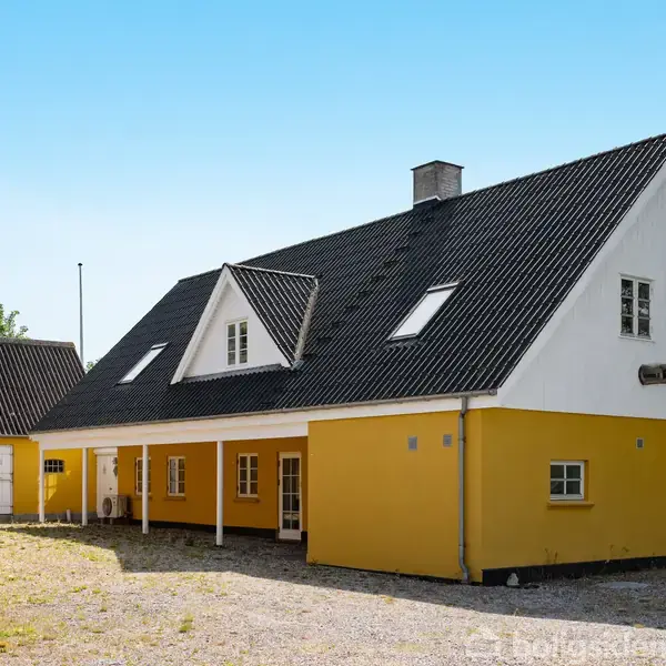 Et gult hus med sort tag står stille i en åben gårdsplads. Huset har flere vinduer, en skorsten og er omgivet af grus og grønne områder under en klar, blå himmel.