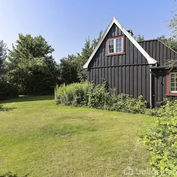 Et sort træhus med røde vindueskanter står omgivet af en grøn have med buske og træer. Huset har en spids tagryg og et vindue på første sal.
