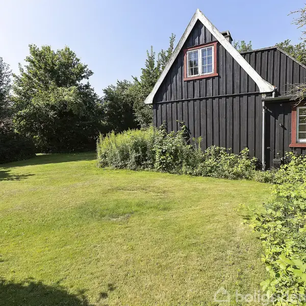 Sort træhus med hvidt gavl pryder haven. Huset står ved frodige buske og græsplæne. Træerne danner en grøn baggrund i det solrige vejr.