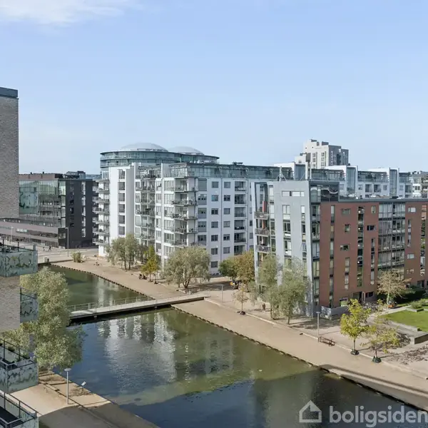 Hvide og grå lejlighedsbygninger med balkoner står langs en kanal med en gangbro. Træer er spredt omkring, og høje bygninger ses i baggrunden under en klar blå himmel.