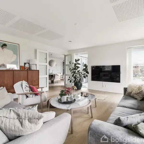 En stue med grå sofaer, et sofabord med planter, og en vægmonteret fladskærm. Baggrunden indeholder en træreol, kunstværk og store vinduer med udsigt til en have.