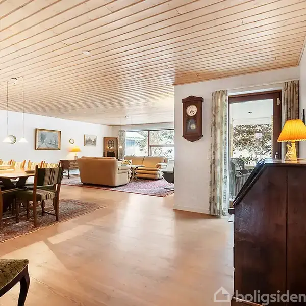 Et rummeligt stueområde med spisebord til otte personer til venstre og en sofagruppe i baggrunden. Trægulv og træloft skaber en varm atmosfære. Udsigt til terrasse gennem glasdør.