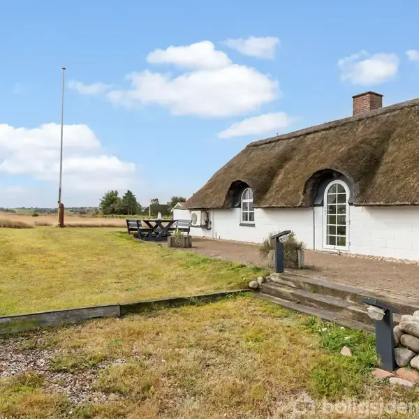 Stråtækt hus står stille på en stor, grøn grund med et bord/bænkesæt. Marken strækker sig mod horisonten under en blå himmel med få skyer.