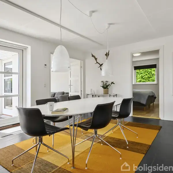 Spisebord med sorte stole står på en orange-gul tæppe i et lyst rum. To lamper hænger over bordet. I baggrunden er hvide vægge og døre til andre rum.