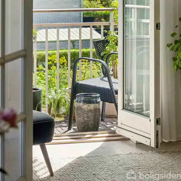 Dør åbner mod en balkon med stol og planter, omgivet af friske, grønne omgivelser. Indendørs er et tæppe og en buket blomster synlige.