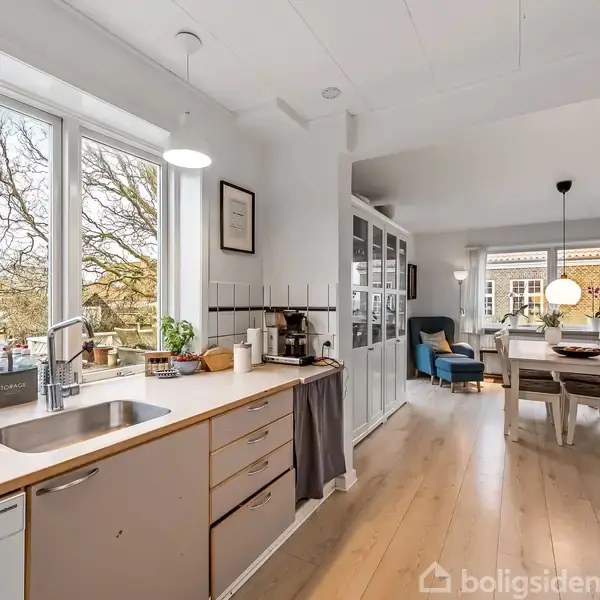 Et lyst køkken med træbordplade og vask. Det åbner op til en spiseplads med et bord og stole, omgivet af store vinduer og naturligt lys.