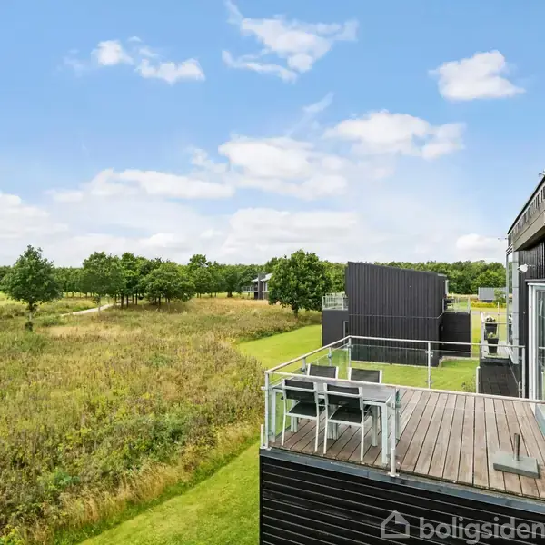 En sort træterrasse med glasgelænder rummer havemøbler; den vender ud mod en åben græsmark med stier og træer, under en blå himmel med få skyer.