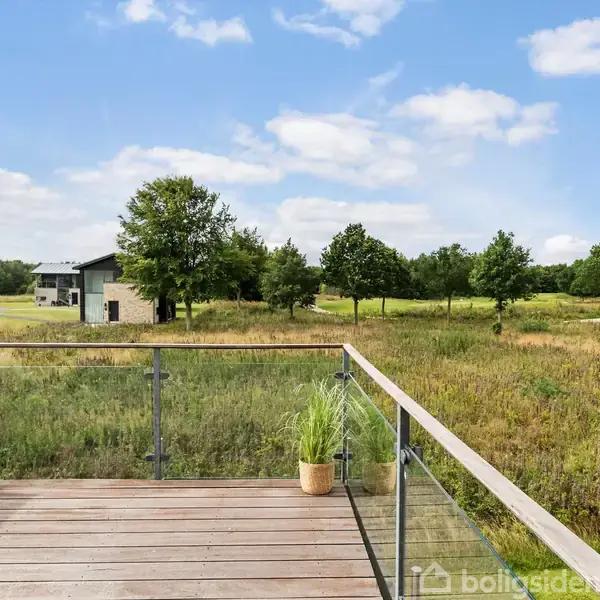 Terrasse med glasrækværk, der fører ud til en åben mark med spredte træer. Et par potteplanter står på rækværket og nogle moderne huse er synlige i baggrunden under en blå himmel.