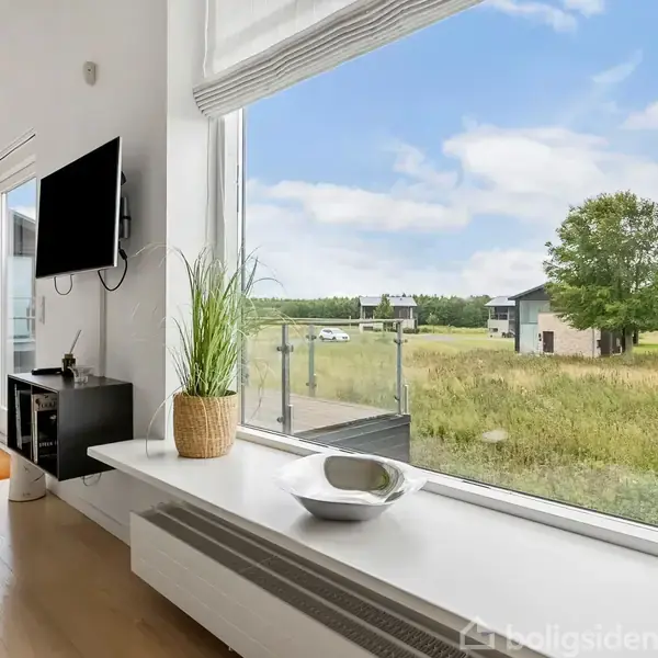 En moderne stue med en stor glasvæg viser udsigt over en grøn mark og træer. Et TV hænger på væggen, og plantepynt står på en hvid bænk.