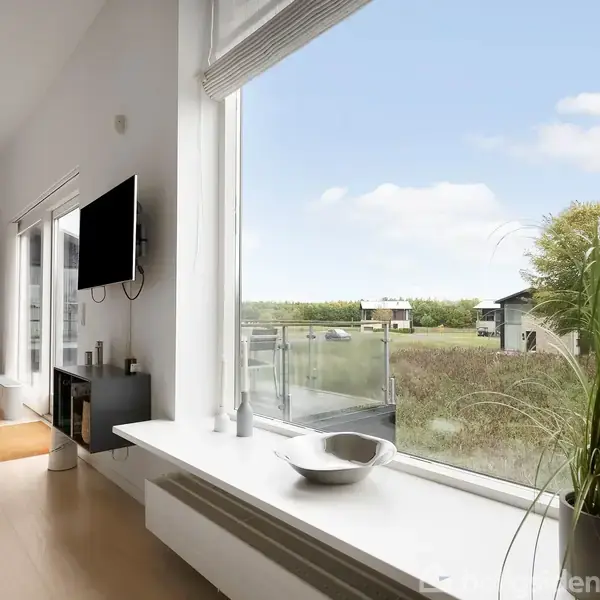 En moderne stue har et stort vindue med udsigt over grønt landskab. Et TV hænger på væggen, og en skål står på vindueskarmen. Der er også en potteplante.
