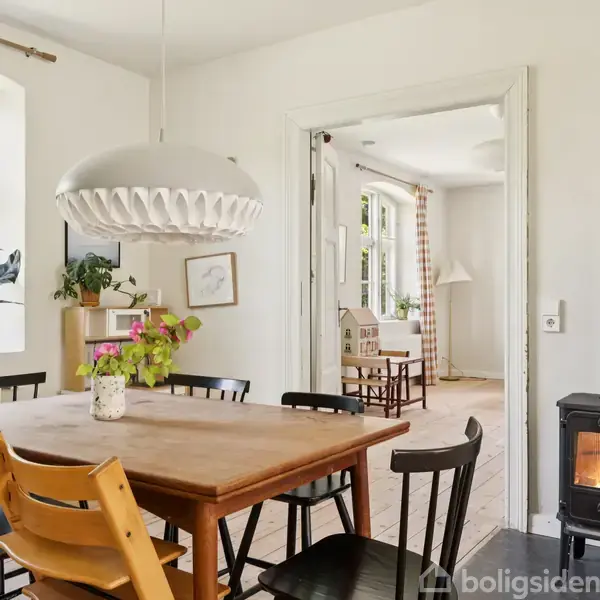 Et træbord omgivet af stole i et lyst rum. En stor, hvid lampe hænger over bordet. En brændeovn står ved siden af, og der er planter på vindueskarmen.