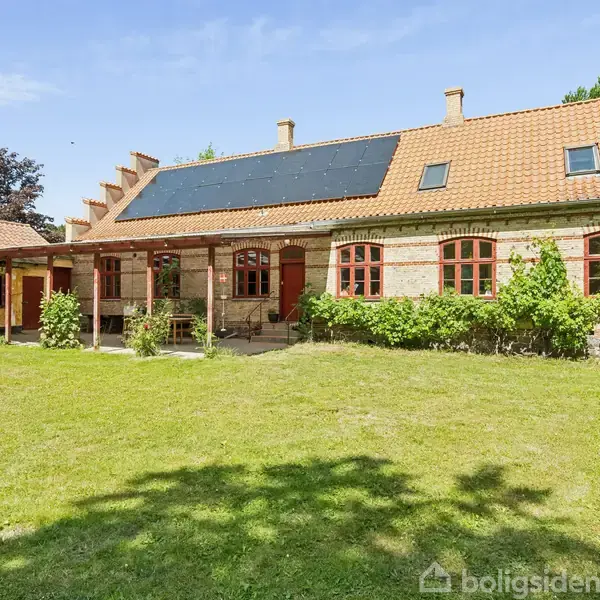 Hus med solpaneler på taget, omgivet af en velplejet have. Bygningen har røde teglsten og røde vinduesrammer, der tilføjer charme. Lyst og frodigt haverum omkring hele huset.