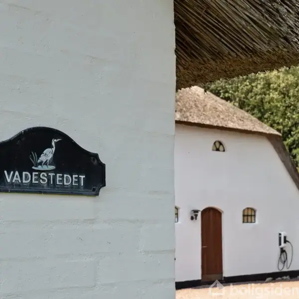 Skilt med teksten "VADESTEDET" hænger på en hvid mur. I baggrunden ses en hvid bygning med stråtag, omgivet af træer og en have.