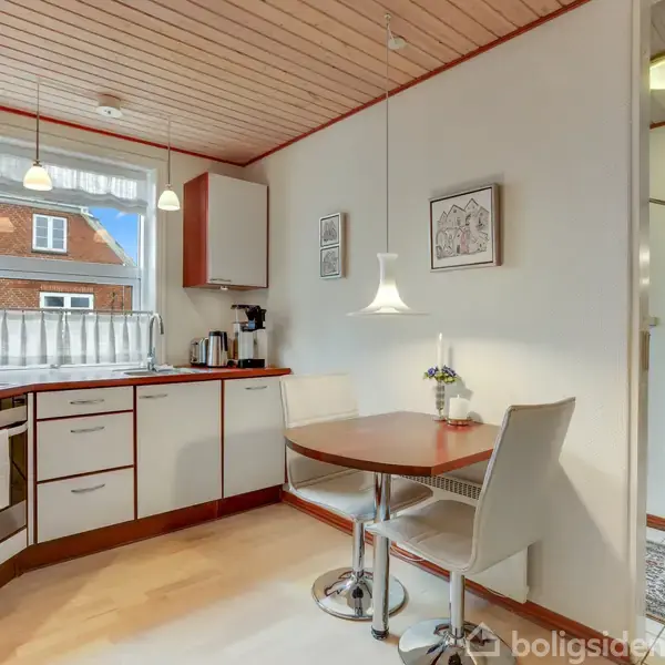 Et køkken med hvide skabe og træ-borde er oplyst af naturligt lys fra et vindue. To moderne stole står ved et rundt spisebord, omgivet af vægdekorationer.