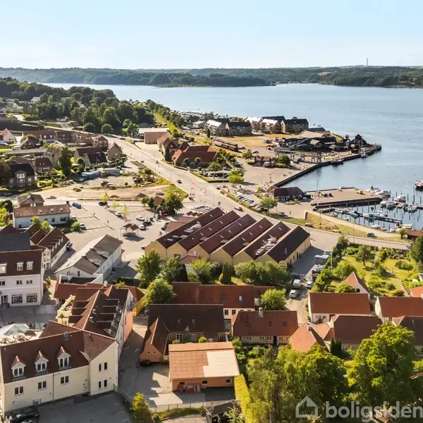 Et kystbyområde med huse og bygninger strækker sig mod en lille havn. Flere både ligger fortøjet ved broerne i en rolig vandkant. Byen er omgivet af frodigt grønt.