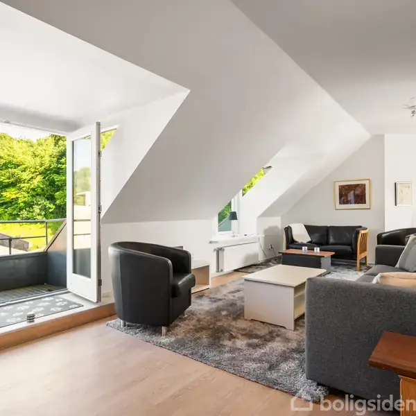 En moderne stue med to grå sofaer og en lænestol omkring et sofabord, vendt mod en åben altandør, der tilbyder udsigt til frodig grønne træer udenfor.
