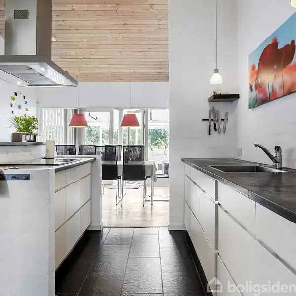 Et moderne køkken med hvide skabe og sort bordplade, en stålvask og ovn; i baggrunden et spisebord med stole og hængende lamper; kunst på væggene.