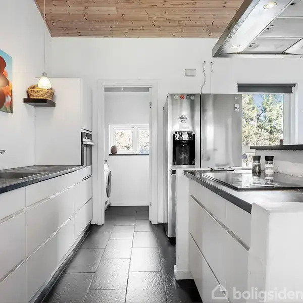 Et moderne køkken har hvide skabe og sorte bordplader, en stålfarvet køleskab og åbent vindue med planter ved siden af. Loftet er træbeklædt, og der hænger et maleri med røde træer.