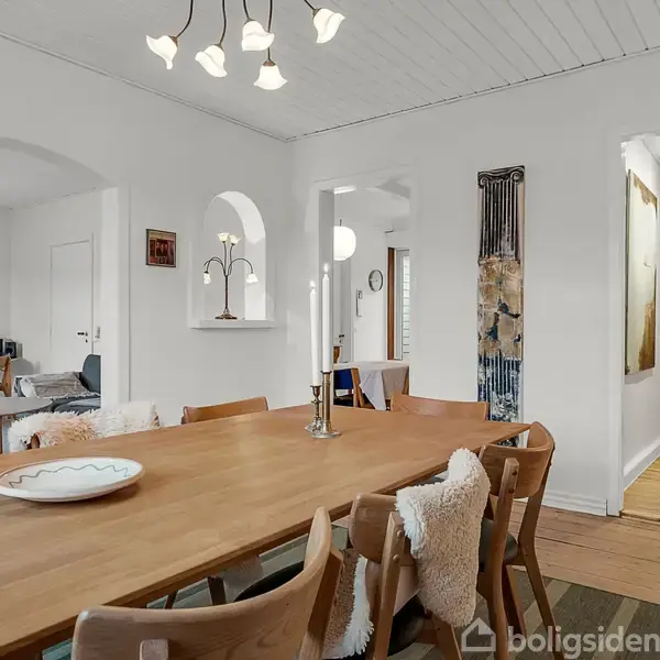 Langt træbord med seks stole og lysestager, placeret i en rummelig spisestue med hvide vægge. Til venstre ses en stue med sofa og skrivebord.