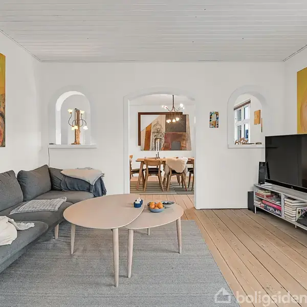 Grå sofa med puder står i stue med træbord foran. Vægkunst og fjernsyn skaber et moderne miljø. Gennem åbningen ses en spisestue med bord og stole.