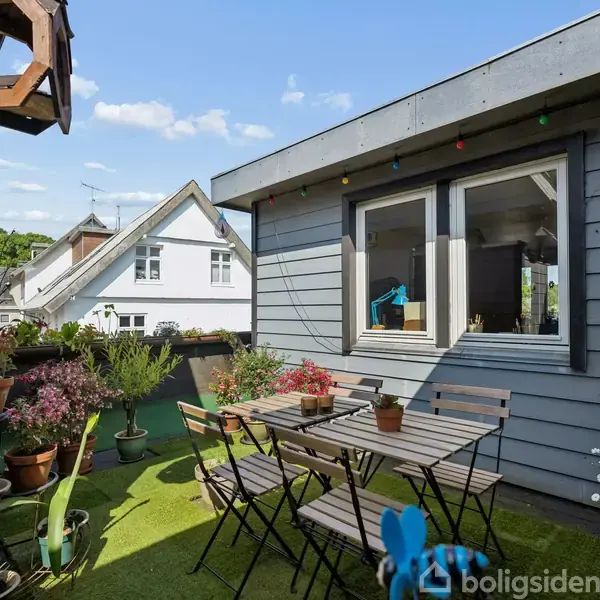 Et havesæt med bord og stole står på en terrasse omringet af blomsterkrukker og planter. Rundt om hænger farvede lyskæder, og der er et fuglehus i træ.