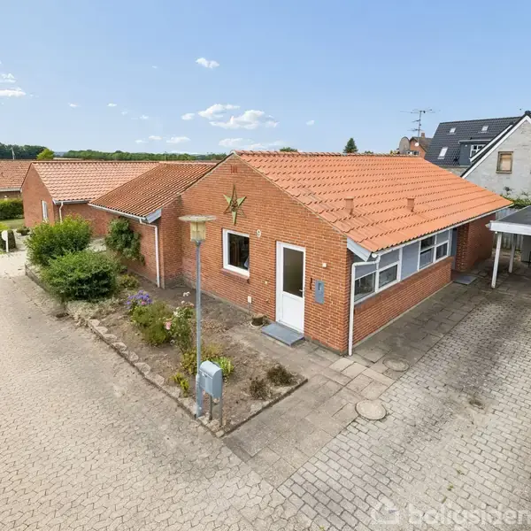 En enkeltstående murstenshus med rødt tegltag står ved en brolagt vej. En lille forhave med buske omgiver huset. Til højre er en carport. Solskinsdag med blå himmel og lette skyer.