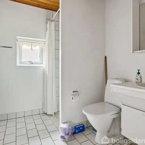 Badeværelse med toilet og hvid håndvask, der står ved en væg. En bruser med hvidt forhæng er i et hjørne. Rummet har hvide fliser og et lille vindue.