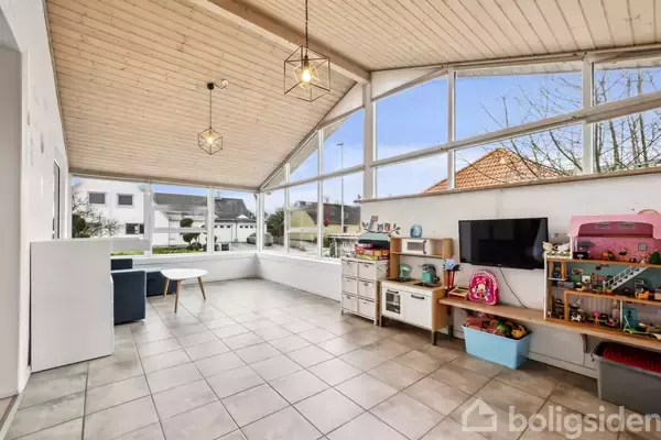 Et lyst rum med legetøjshylder, et tv og borde arrangeret mod store vinduer. Rummet har flisegulv, træloft og udsigt til et hus og have udenfor.