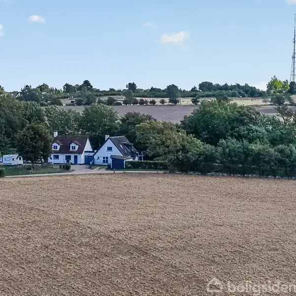 Et hvidt landhus med rødt tag er omkranset af træer ved en stor, åben mark under en blå himmel med spredte skyer. I baggrunden ses en telekommunikationsmast.