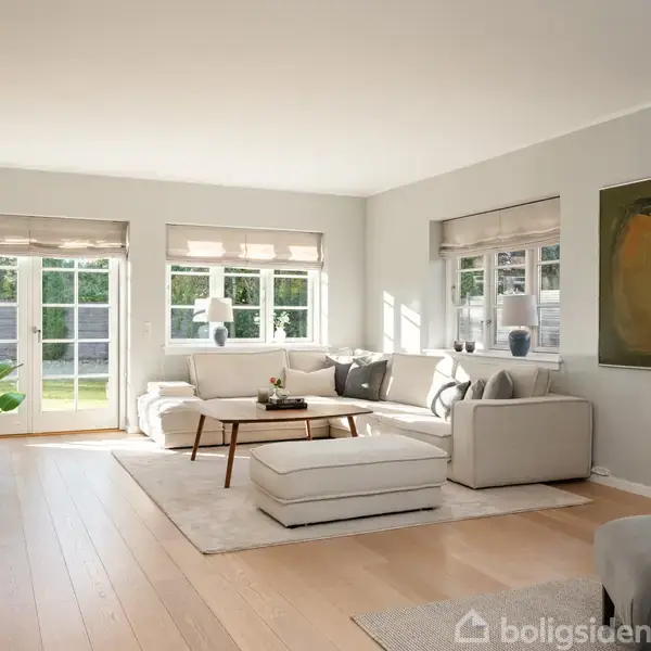 En stor, lys stue med en grå sofa arrangeret omkring et sofabord. Stuen har store vinduer og en glasdør, der åbner ud til en grøn have.