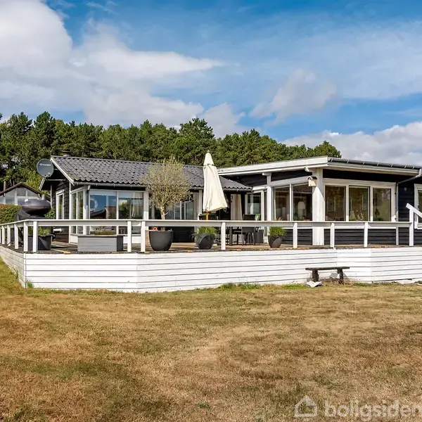 Sommerhus med terrasse, der er fyldt med planter og en parasol, beliggende i natur omgivet af grønt græs og skov. Terrasseafskærmningen er hvid og fremtrædende.
