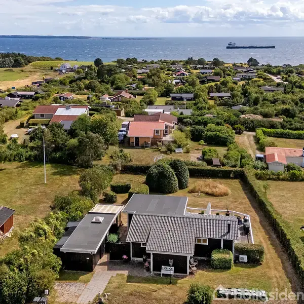 Huse ligger i et grønt område ved kysten med udsigt over havet. En båd sejler forbi i baggrunden. Området er fyldt med træer og markerede haver.