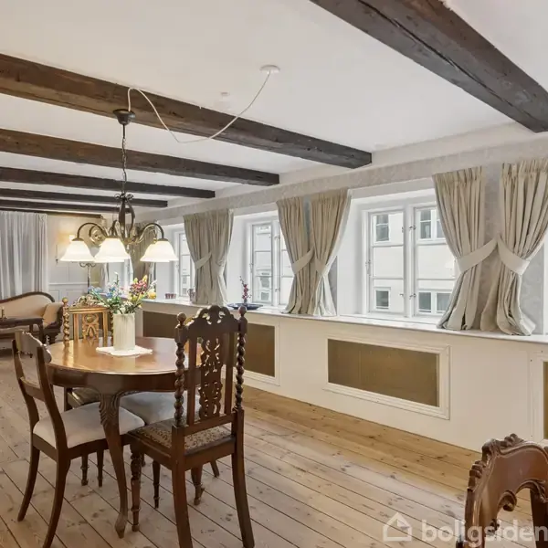 Et spisebord med fire stole står på et trægulv i en stue. Loftsbjælker ses ovenover, og store vinduer med gardiner fylder rummet med lys.