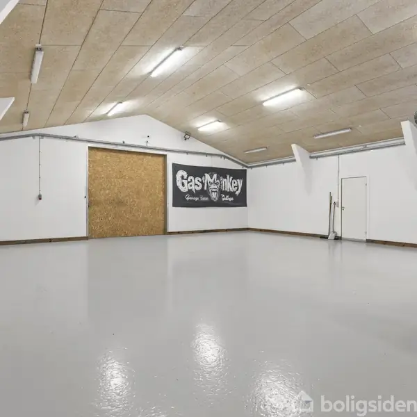 Garage med to biler, den ene hævet på en lift. Væggene er hvide, og loftet har indbyggede spotlys. Et skilt med teksten "Gas Monkey" hænger på væggen.
