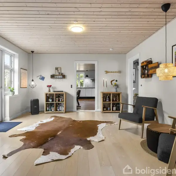 Stue med lyst trægulv og -loft, ko-skindstæppe, to lænestole, bord, hylder, og en pendellampe. Store vinduer og dør fører til en terrasse. En plakat hænger på væggen.