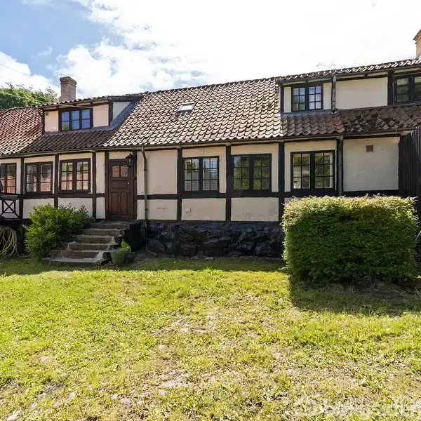 Et bindingsværkshus med rødt tegltag og flere skorstene står stille. Foran huset er en velplejet græsplæne og en busk. Det er en solrig dag med blå himmel.