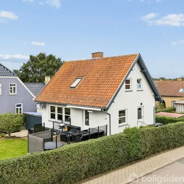 Et hvidt hus med rødt tegltag står frit. Terrassen har havemøbler. Omgivet af en klippet hæk og en græsplæne i en villavejsomgivelse med andre huse.
