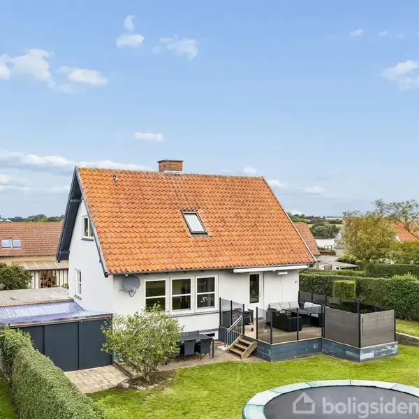 Hus med rød tag og hvid facade, omgivet af en grøn have. En træterrasse med møbler ses ved siden af huset. Trampolin placeret på græsplænen. Skyfri blå himmel i baggrunden.