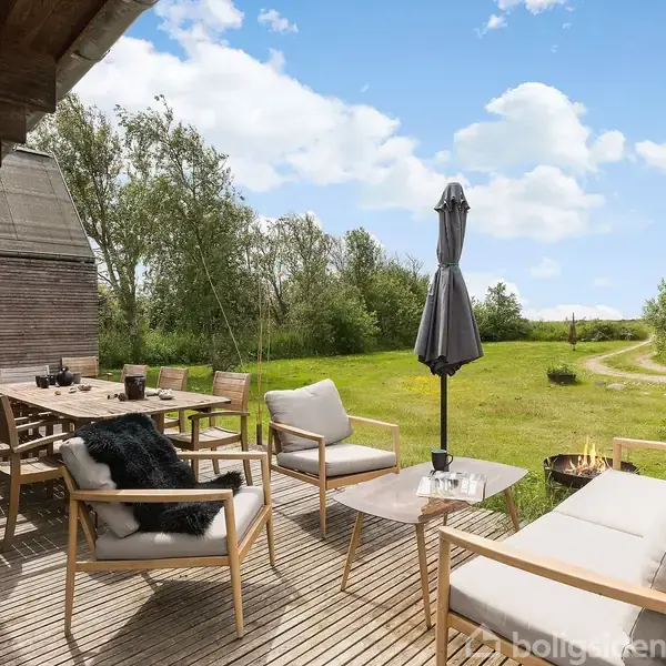 Terrasseområde med træmøbler og haveparasol, placeret ved siden af et træhus. Møblerne inkluderer en lang spisebordssæt og lounge-stole. Græsplæne og træer omkranser området under en klar himmel.