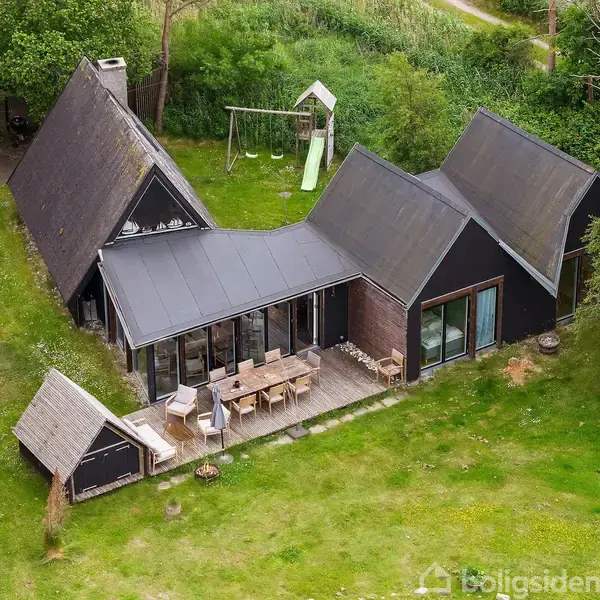 Hus med skrå tage udendørs sidder i en grøn have med en terrasse fyldt med havemøbler. I baggrunden er der en gyngestativ og skydestige omgivet af træer og buske.