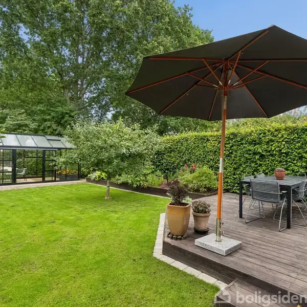 Stor parasol står på træterrasse med havemøbler, omgivet af velplejet have med grøn græsplæne og buske. I baggrunden ses et drivhus foran en række træer.