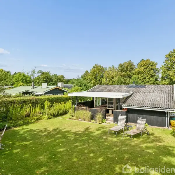 Et sommerhus med et mørkt tag står midt i en frodig have. To havestole er placeret foran huset, og et havebord med stole ses til venstre, omgivet af grønne buske og træer.