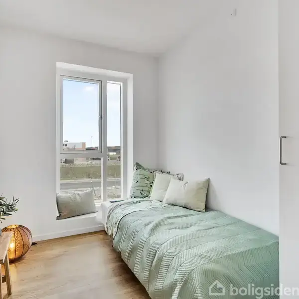 Et lille værelse med en enkeltseng dækket af et grønt tæppe, står ved siden af et vindue. På en træbænk ved siden af er der en plante og en lampe.