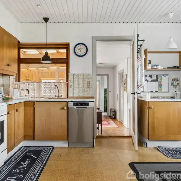 Et køkken med brune skabe, en ovn og en opvaskemaskine. Der er en vægudsmykning med en blå ur. Trægulv og fliser på væggene. Tæpper mærket "My Kitchen".